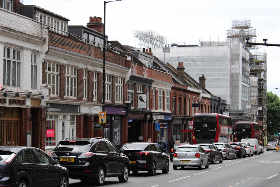 The image shows a busy street scene on Ballards Lane with a row of multi-story terraced buildings featuring brick facades, large windows, and some with decorative white trim, under partly cloudy skies. Along the pavement, there are parked cars including sedans and hatchbacks, with a black Mercedes-Benz and a silver vehicle closest to the camera. Two red double-decker buses, numbered 343 and 42, are visible in motion along the street, indicating local public transport activity. Pedestrians are walking along the sidewalk, some near small shops and businesses that have signs and window displays. On the right side of the street, a building under renovation is covered in scaffolding and white protective sheeting, suggesting ongoing construction or refurbishment. The street scene captures an active urban environment with a mix of residential and commercial buildings, typical of a central London neighbourhood, and provides context relevant to house removals and moving services by illustrating the urban infrastructure and transport logistics involved in a home relocation or furniture transport process facilitated by North Finchley Removals.