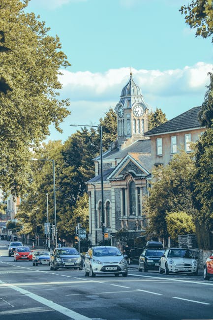 A street scene with multiple parked cars along the curb, including a white van positioned in front of a large, historic building with a clock tower. The building features architectural details such as arched windows and decorative stonework, surrounded by lush green trees that line the street. The sky is bright and partly cloudy, casting natural daylight over the scene. This setting depicts an urban environment suitable for house removals or furniture transport services, as provided by North Finchley Removals. The scene suggests the process of home relocation involving the loading or unloading of household items, with the clock tower serving as a notable landmark in the area. The street is calm, with trees providing shade, and the overall atmosphere emphasizes a typical day in a residential and commercial vicinity where professional removals might take place.