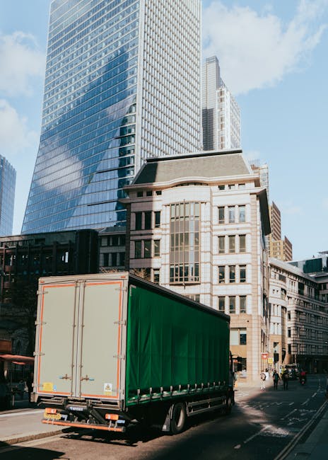 A large green delivery truck parked on a city street with tall modern office buildings and older architectural structures in the background. The truck is positioned with its rear doors closed, ready f