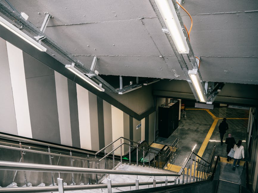 Inside a stairwell at Finchley Central station, a metallic staircase with a safety railing is visible, descending from an upper level with a grey tiled ceiling fitted with linear fluorescent lights. The walls are decorated with vertical grey and beige stripes, and a black cabinet is positioned near the bottom of the stairs. On the station's pavement area outside the stairs, three individuals are walking away from the camera, carrying bags and items, and moving towards the station's exit. The pavement features yellow tactile paving strips for accessibility, with a sloped pathway leading toward the exit, which is visible in the background. This scene captures the typical environment associated with house removals and furniture transport planning, illustrating the internal and external movement logistics involved in a home relocation facilitated by North Finchley Removals.