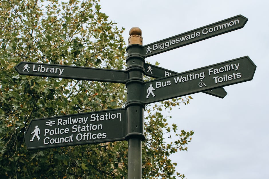 A black multi-directional street signpost situated outdoors against a background of leafy green trees and a cloudy sky. The signposts indicate directions to various local amenities including Biggleswade Common, a library, a railway station, a police station, council offices, a bus waiting facility, and toilets, with symbols for pedestrians and wheelchair accessibility. The signpost is part of a typical urban setting, helping guide residents and visitors during home relocation or furniture transport arrangements facilitated by North Finchley Removals, as detailed on the Finchley Central station access guide page.