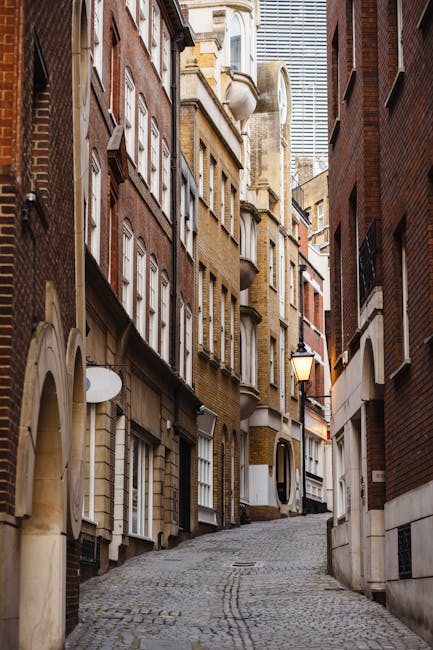 A narrow urban street scene during daytime, flanked by tall, closely spaced buildings with residential and commercial signage, including a hotel sign on the left. The street is paved with cobblestones