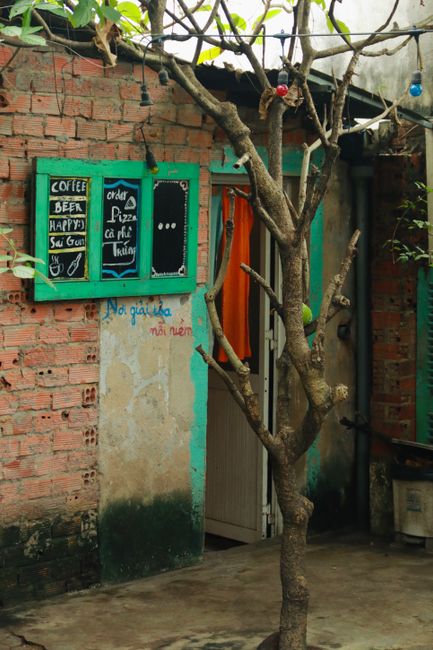 A photograph showing the exterior of a building with a red brick wall and a small, partially painted concrete section. Attached to the wall are three black chalkboards with colourful chalk writing advertising coffee, beer, pizza, and other items, with some words in Vietnamese. In front of the wall, there is a small, leafless tree with multiple branches extending upward and outward, casting shadows on the wall. To the right, a partially open door with an orange curtain is visible, leading into the building interior. A metal gutter runs along the roof edge above, with a black pipe on the wall nearby. The ground is concrete with some dark patches, indicating weathering. The scene is illuminated by natural daylight, creating shadows and highlights on the textured surfaces. The surroundings suggest a modest, urban environment potentially involved in home relocation or small move activities, relevant to the services provided by North Finchley Removals.