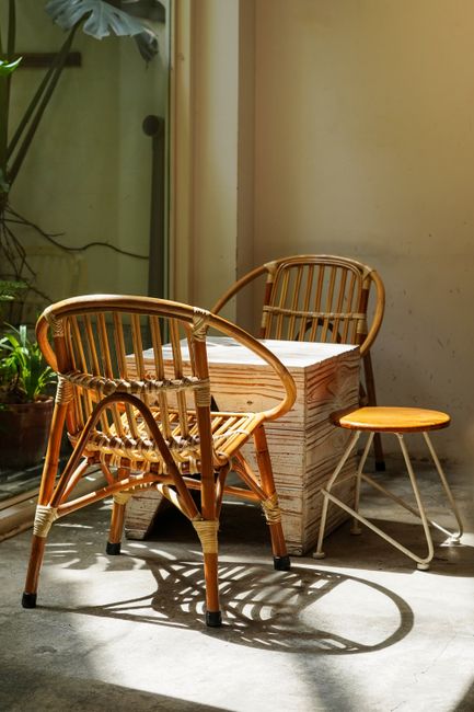 A corner of an indoor space featuring two wicker chairs with rounded backs and armrests, placed near a light-colored wooden table with a rectangular shape and visible wood grain, accompanied by a small round wooden stool with a metal frame. The chairs and stool are positioned on a concrete floor, with sunlight casting shadows from the furniture. In the background, there is a potted plant with green leaves, a section of a wall, and a glass window or door, indicating a room likely used for casual seating or relaxation during a home relocation or furniture transport process. The setting appears tidy and well-lit, with natural light enhancing the textures of the furniture and surroundings, and the scene subtly aligns with house removals or packing activities supported by North Finchley Removals.